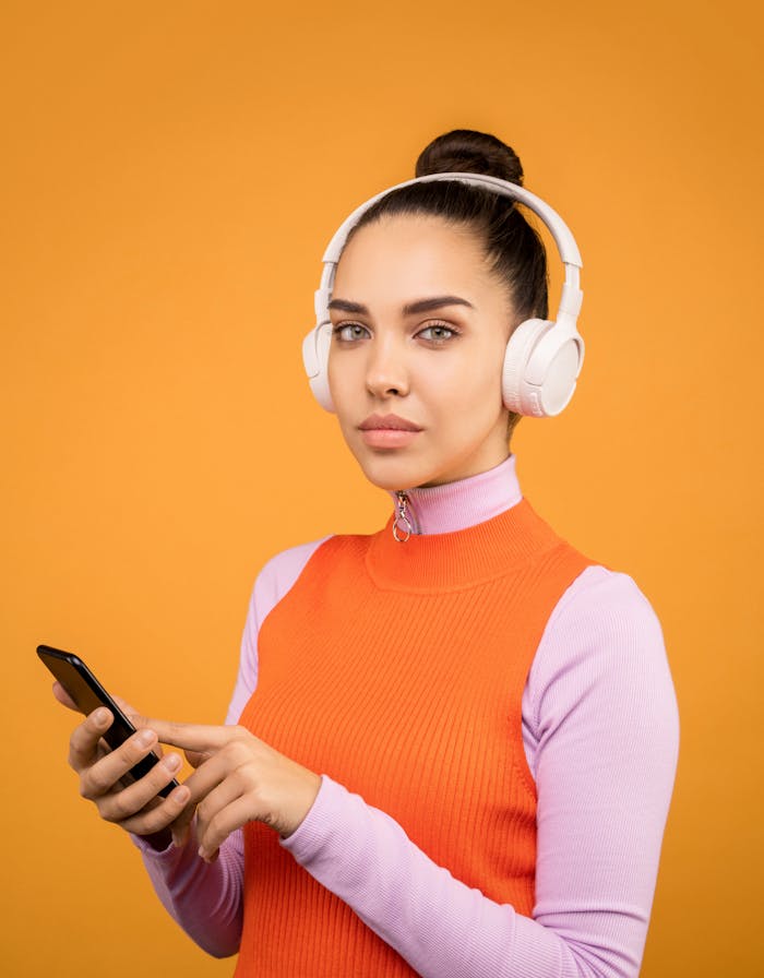 A young woman listens to music on wireless headphones while using a smartphone against an orange background.